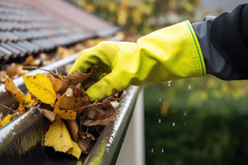 Hand in glove cleaning wet autumn leaves from a roof gutter during maintenance